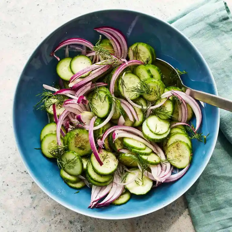 A top-down view of a blue bowl filled with a fresh cucumber salad made of sliced cucumbers, thinly sliced red onions, and…