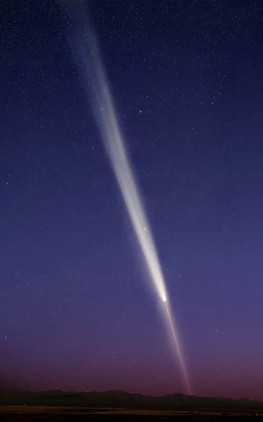 A night sky photo shows a bright comet with a long, glowing tail streaking diagonally across a starry purple and blue sky…