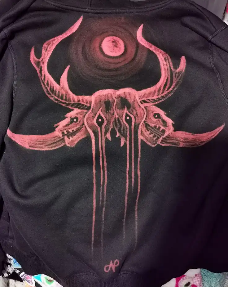 A black hoodie features a large, hand-drawn pink graphic of a demonic skull with three faces, curved horns, and a swirling…