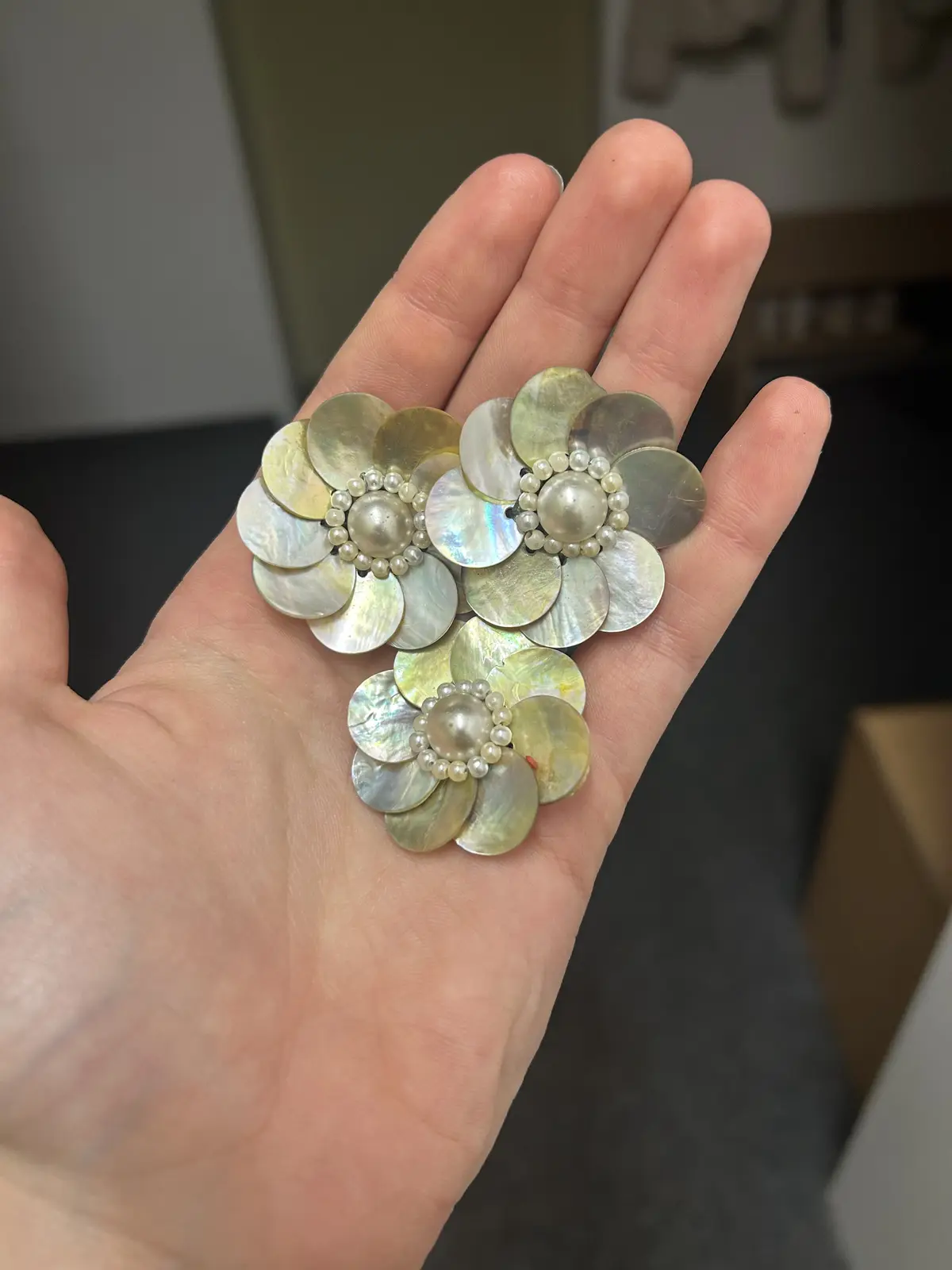 A hand holds a decorative brooch made of iridescent mother-of-pearl petals arranged in three flower shapes, each with a…