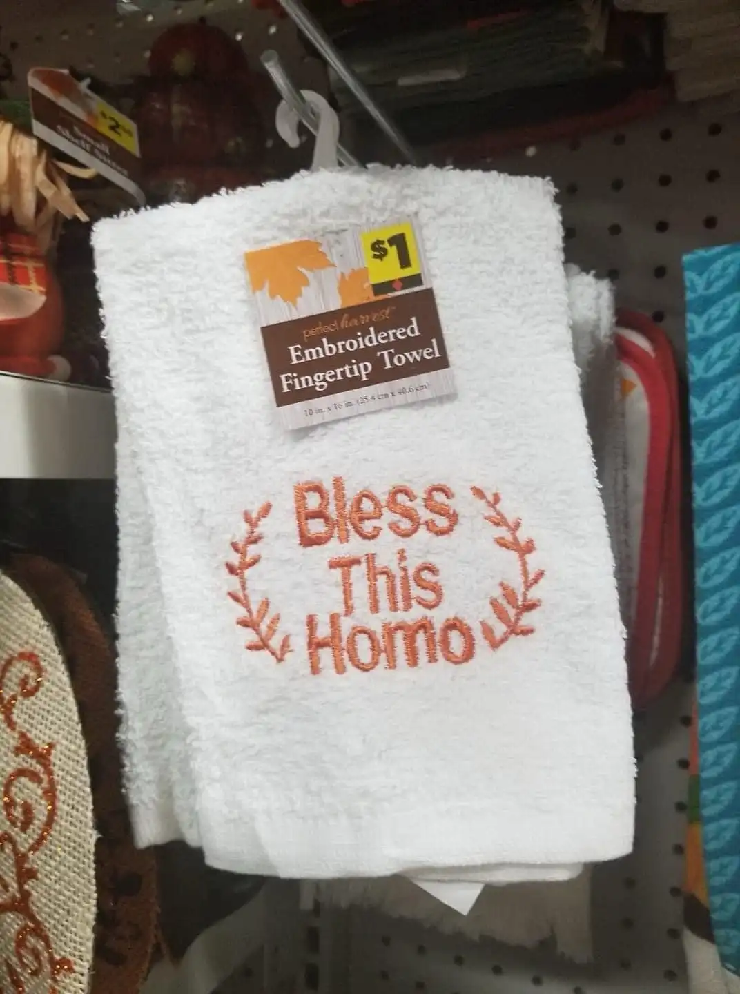 A white embroidered fingertip towel, hanging in a store aisle, has the phrase 'Bless This Homo' stitched in orange thread…