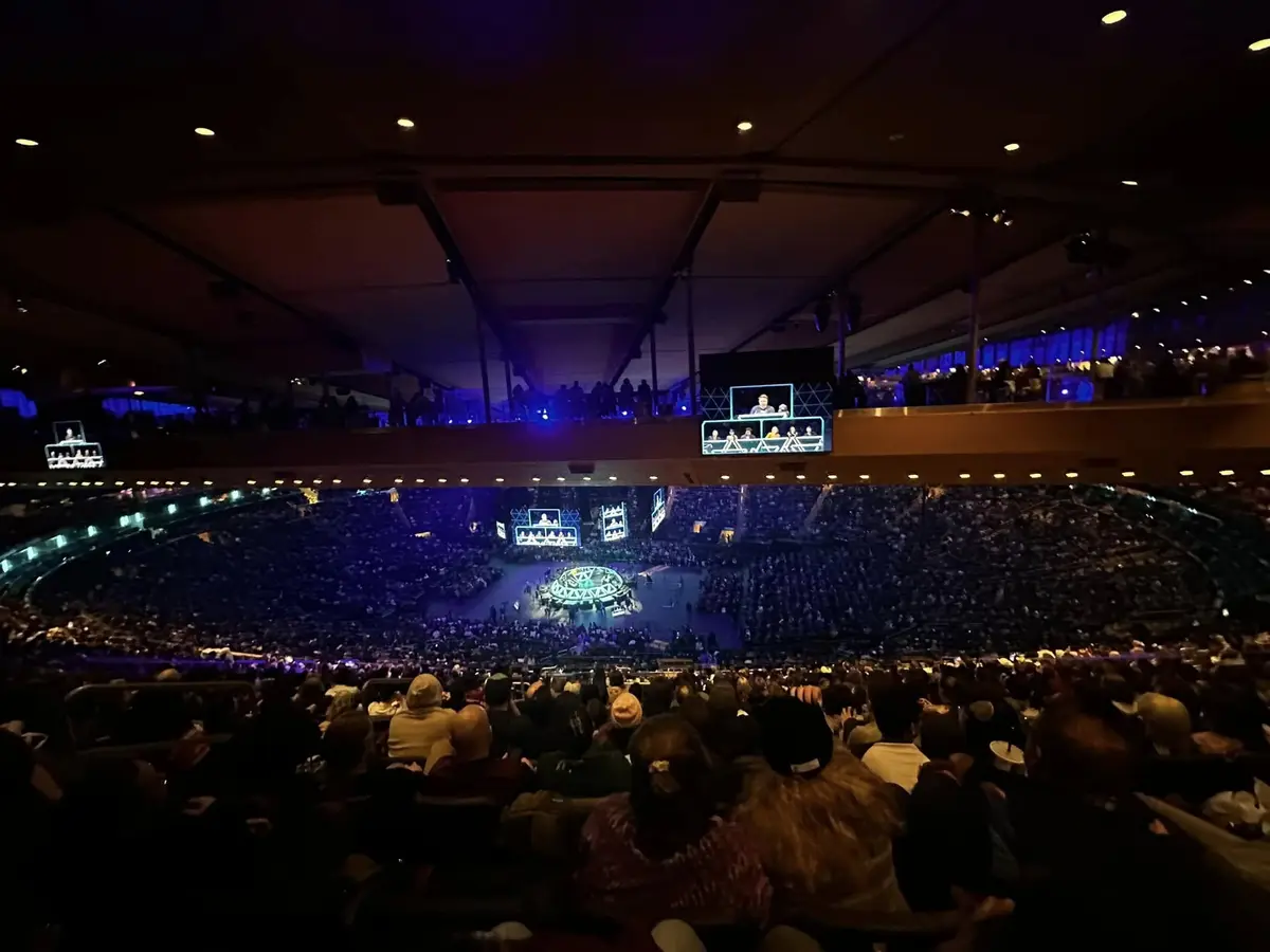 A wide-angle photo from the upper tier of a packed, dimly lit arena during a large-scale event, showing a central stage with…