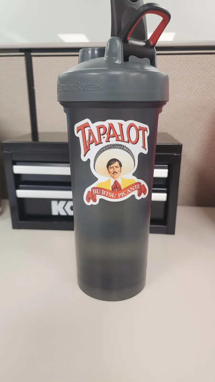 A black BlenderBottle shaker cup sits on a desk, featuring a sticker parodying the Tapalot brand with a man in a sombrero…