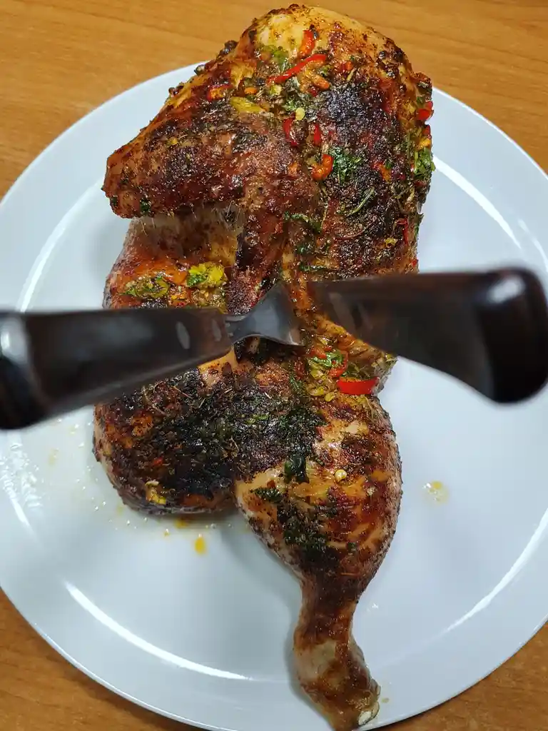 A roasted chicken with a dark, charred skin and visible herbs and red pepper flakes sits on a white plate. A knife and fork…