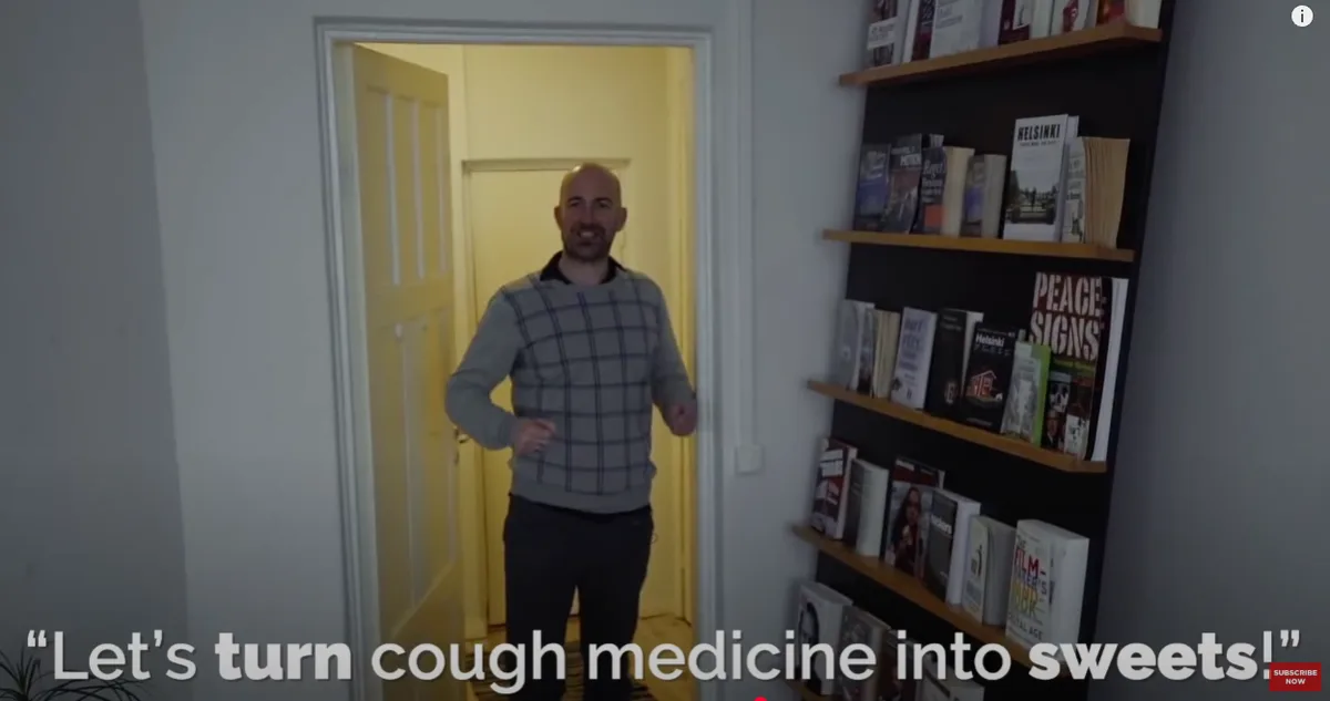 A bald man in a grey plaid sweater stands in a doorway smiling broadly, with a bookshelf visible to his right. The overlaid…