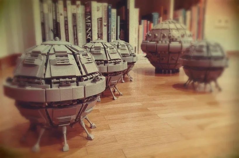 A row of gray Lego Death Star models sits on a wooden floor, with a bookshelf in the background. The foreground model is in…