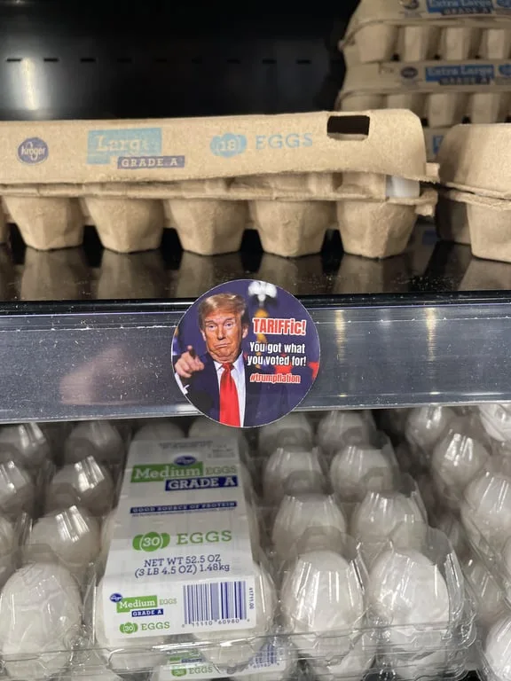 A circular sticker featuring Donald Trump pointing angrily is stuck on a supermarket shelf divider. The sticker has red text…