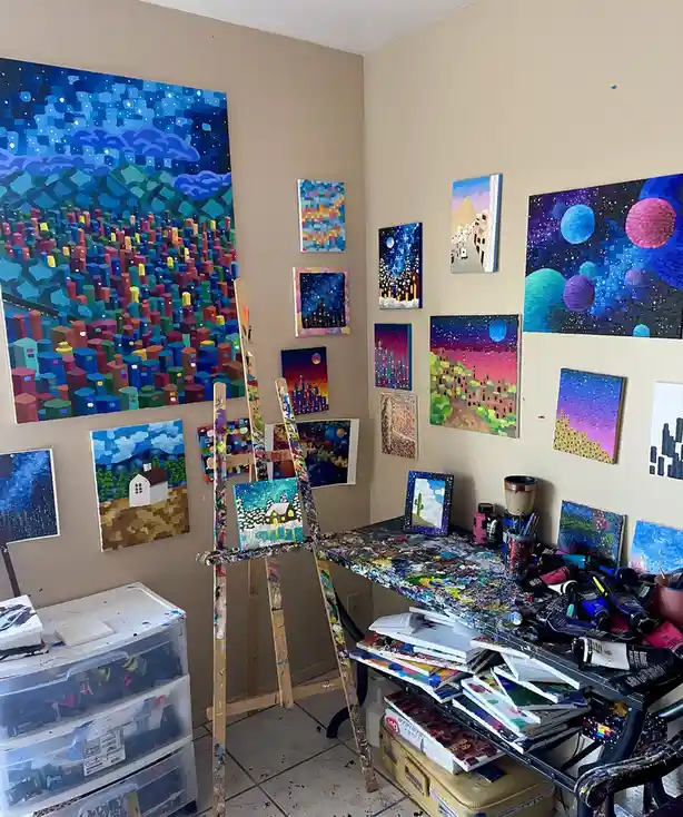 A cluttered artist's studio filled with colorful, pixelated-style paintings on the walls and a messy desk covered in paint…