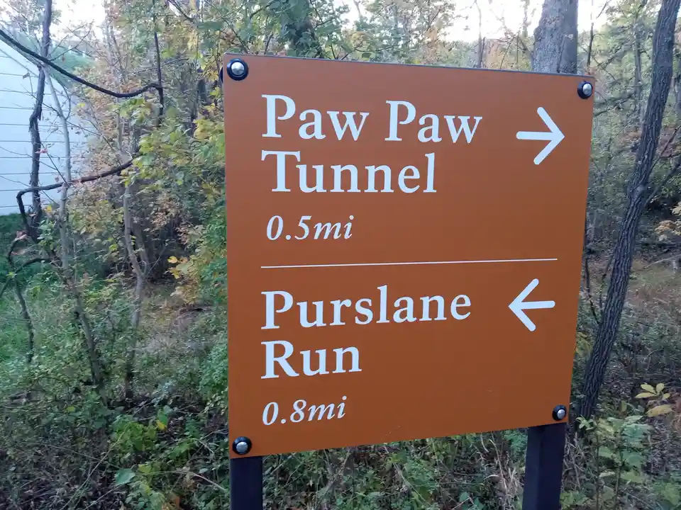 An orange trail sign in a wooded area points right to 'Paw Paw Tunnel' (0.5mi) and left to 'Purslane Run' (0.8mi). The sign…