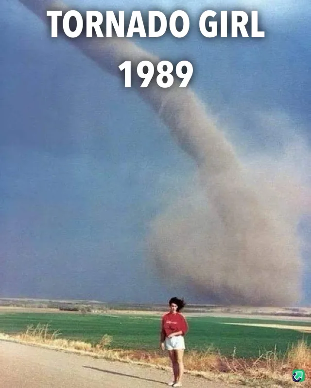 A vintage photograph shows a young woman standing calmly on a rural road, with a large, visible tornado swirling in the…