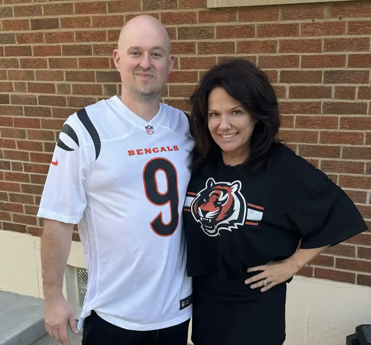 A man and woman stand together in front of a brick wall, both wearing Cincinnati Bengals apparel. The man wears a white…