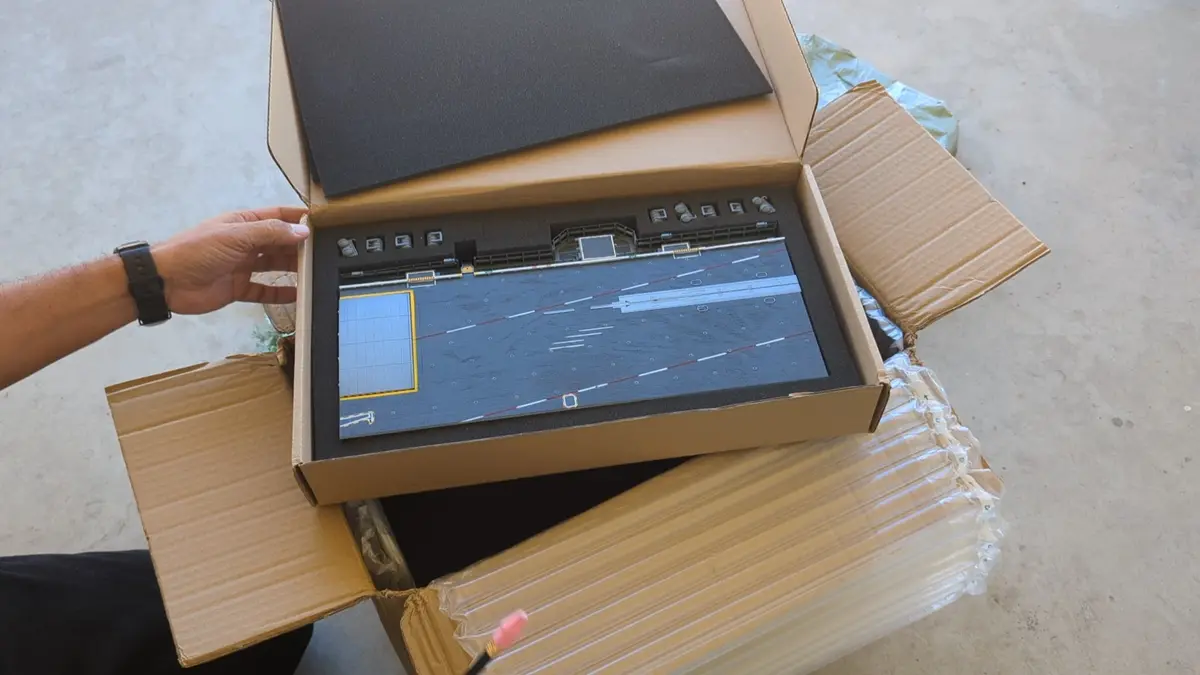 A person's hand opens a large cardboard box revealing a detailed miniature airport runway model with taxiways, markings, and…