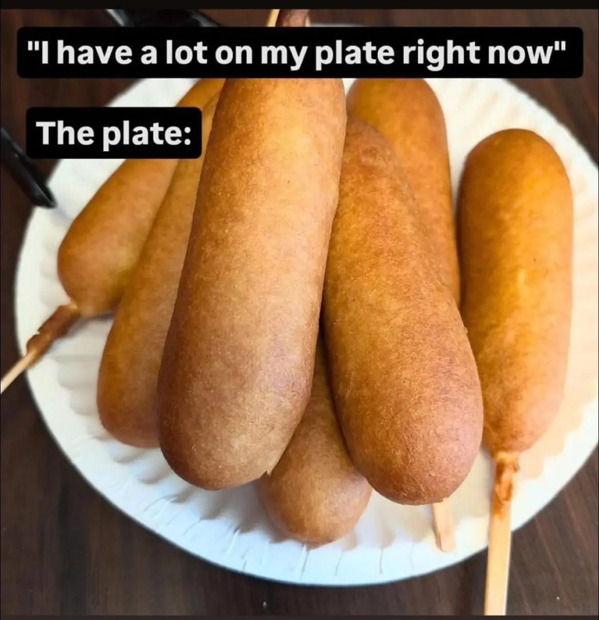 A white paper plate is piled high with numerous golden-brown corn dogs on wooden sticks, visually representing an…