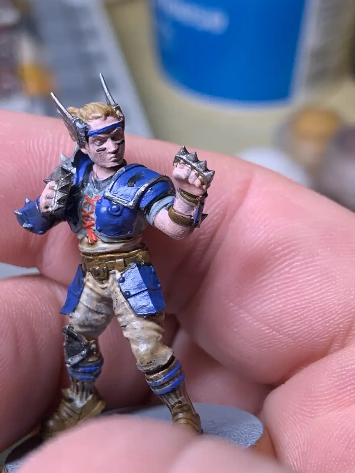 A close-up photo shows a hand holding a small, painted fantasy miniature figure wearing blue armor and spiked gauntlets. The…
