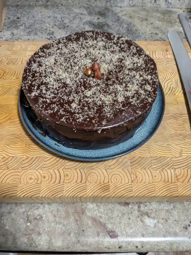 A round chocolate cake with a glossy glaze, topped with shredded coconut and three whole hazelnuts, sits on a blue plate…