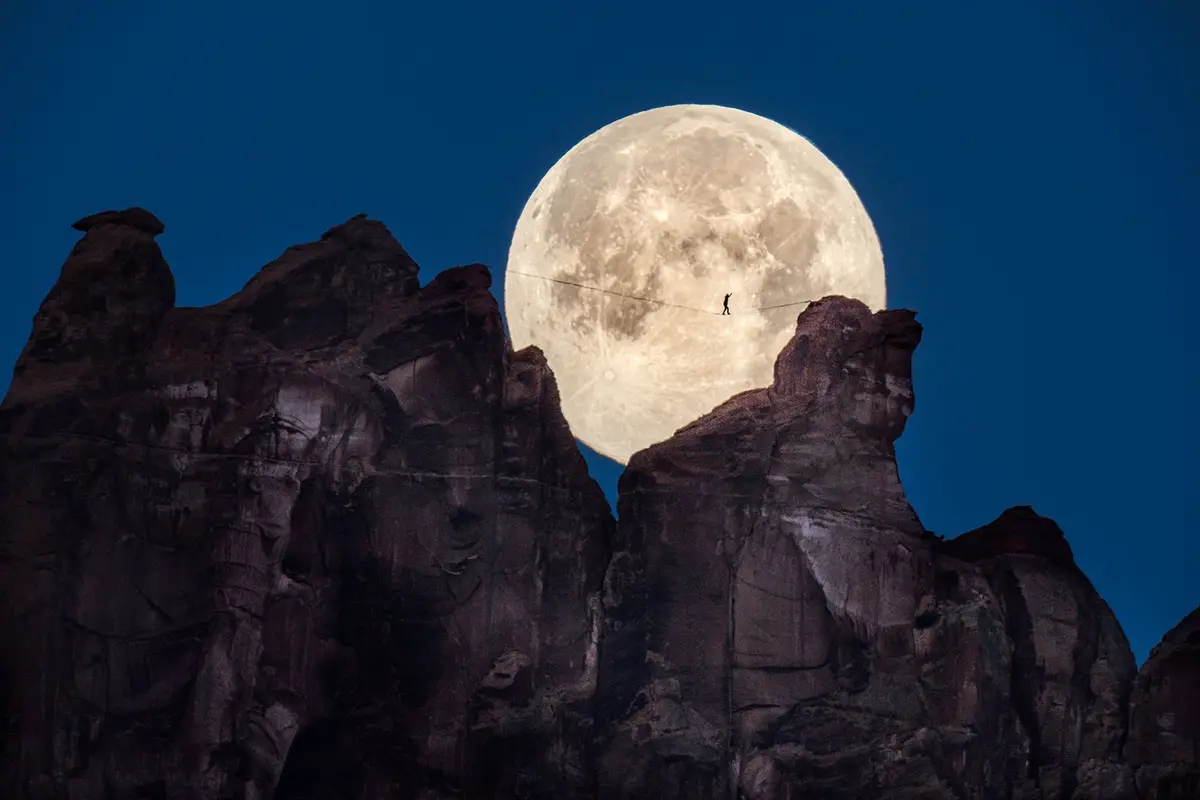 A person is silhouetted walking a tightrope stretched between two large rock formations, with a massive, detailed full moon…