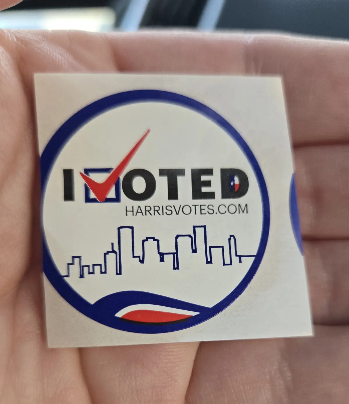 A hand holds a square white sticker with a blue circular border. Inside, the text 'I VOTED' is displayed, with the 'I'…
