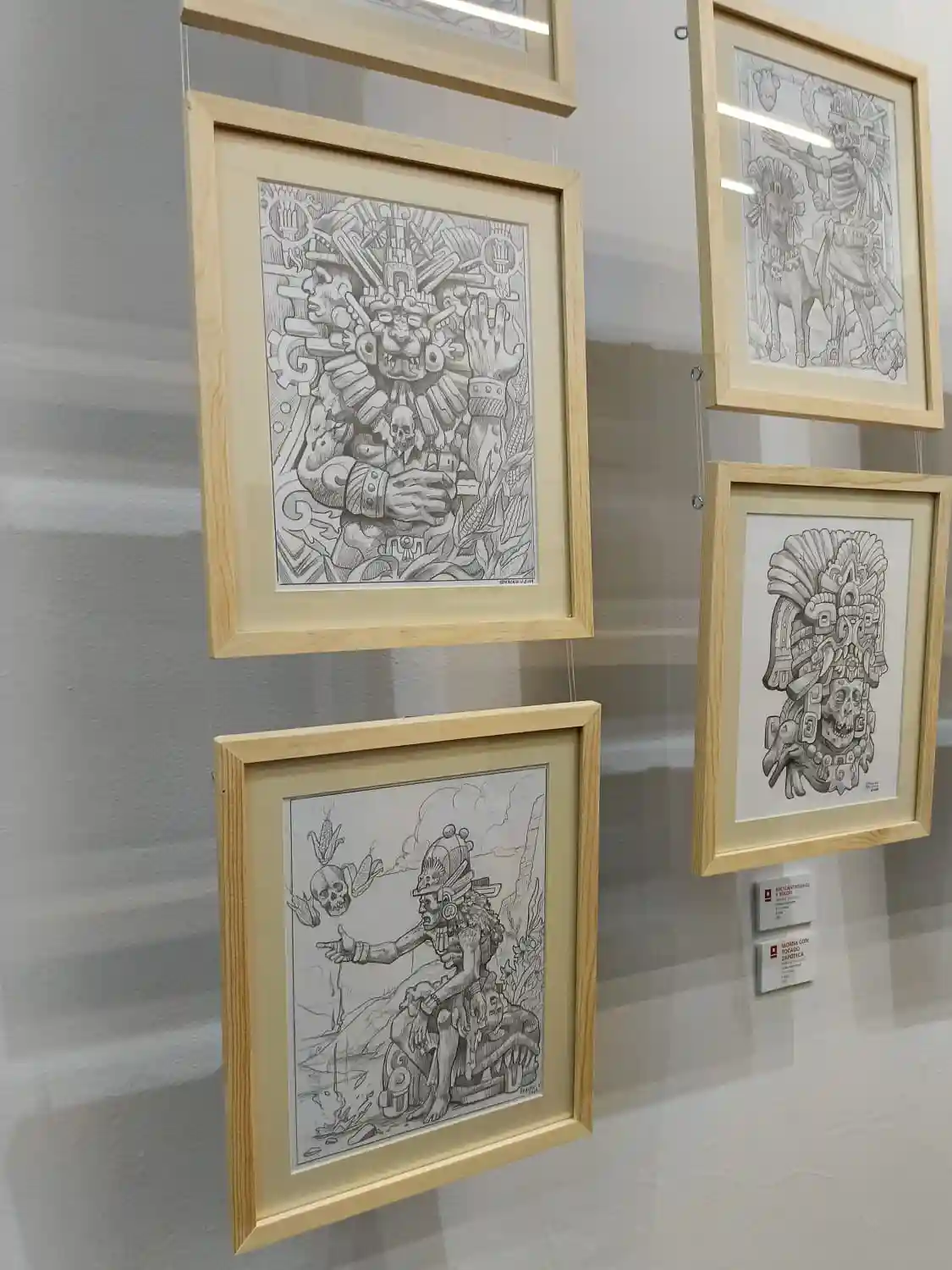 Four framed black-and-white drawings of Mesoamerican deities and mythological figures hang on a gallery wall. The artwork…