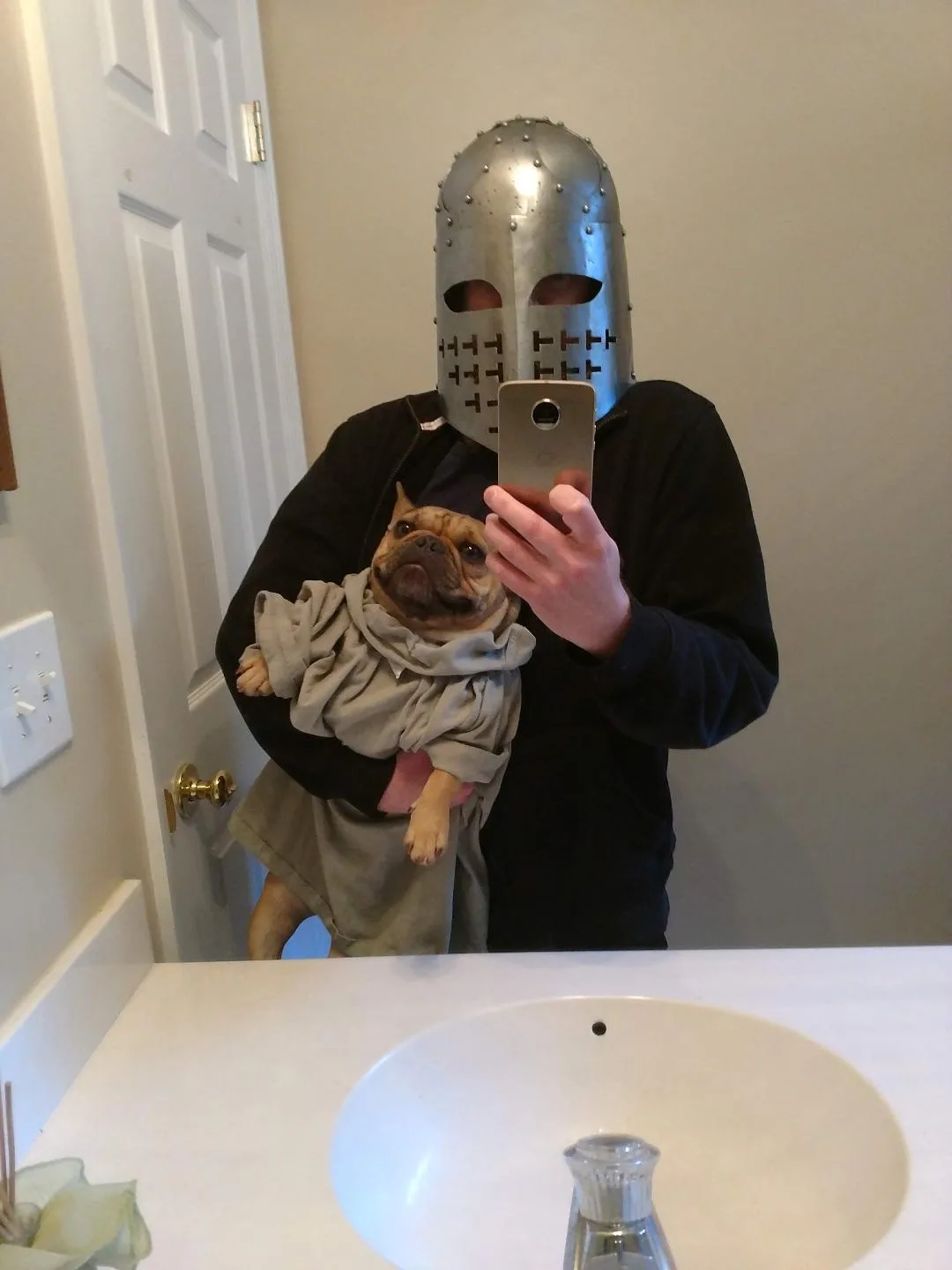 A person wearing a full metal knight's helmet takes a mirror selfie in a bathroom while holding a French bulldog dressed in…