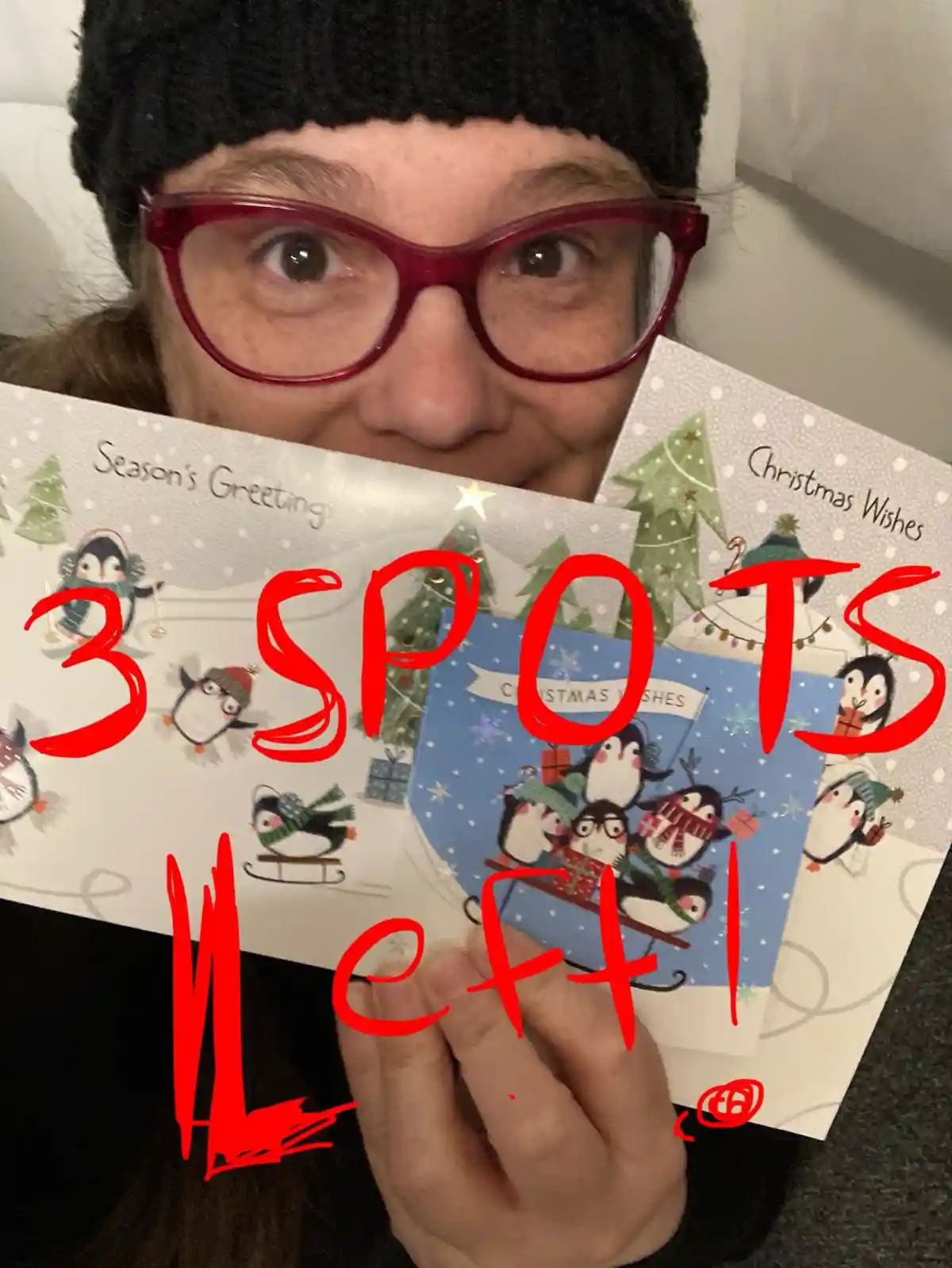 A person wearing red glasses and a black beanie holds up several Christmas cards featuring penguins and holiday scenes…