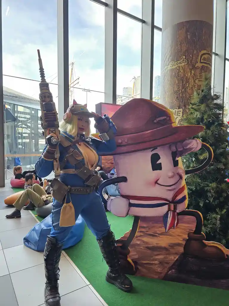 A person in a Fallout-style cosplay, holding a large weapon and wearing a 'Vault 30' cap, poses next to a large cardboard…