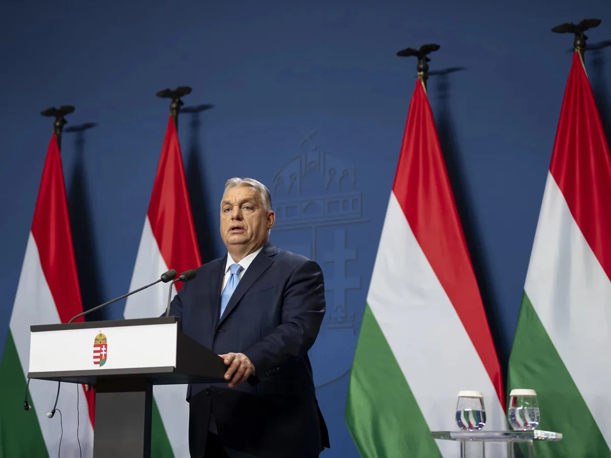 A man in a dark suit and light blue tie stands at a podium with the Hungarian coat of arms, speaking into microphones…