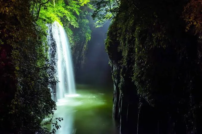 A serene, misty waterfall cascades down a rocky cliff into a calm, green pool, framed by dense, dark foliage on either side…