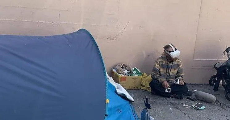 A person sits on the ground next to a blue tent and a cardboard box, wearing a VR headset while holding a controller. The…