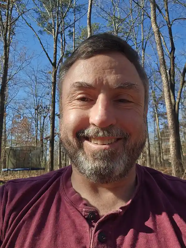 A man with a graying beard and mustache smiles broadly at the camera while wearing a maroon henley shirt, standing outdoors…