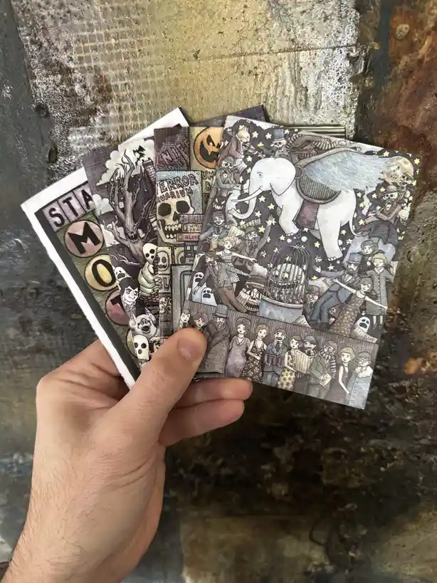 A hand holds a fanned-out set of five illustrated cards against a rusty metal background. Each card features a different…