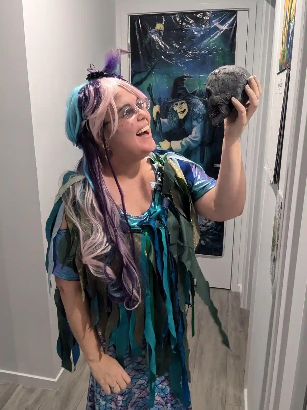 A person in a colorful mermaid costume with pink, blue, and purple hair holds up a skull while smiling broadly. Behind them…