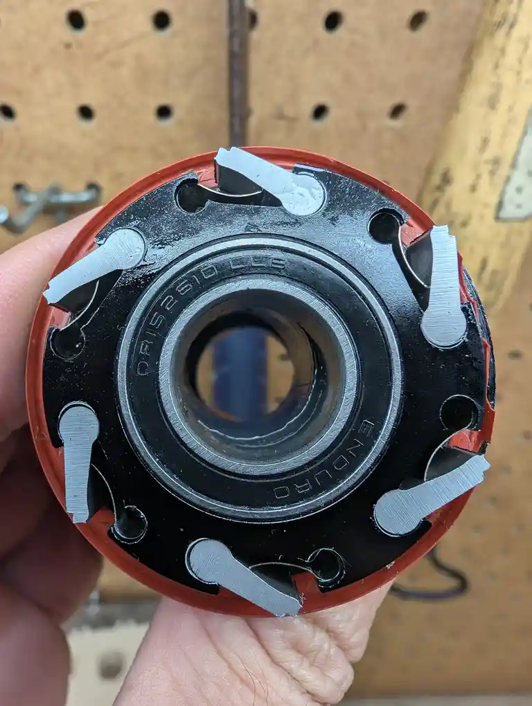 A hand holds a circular router bit with a red outer casing and black inner body, showing multiple broken silver cutting…