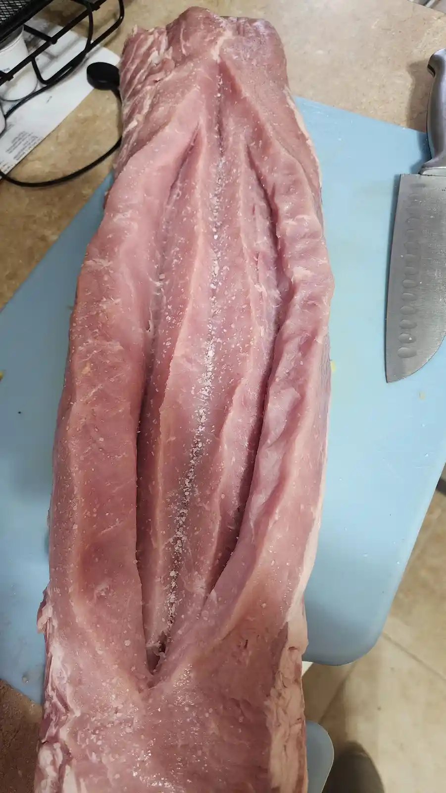 A large, raw piece of meat, likely pork ribs, sits on a blue cutting board with salt sprinkled on it, next to a large chef's…
