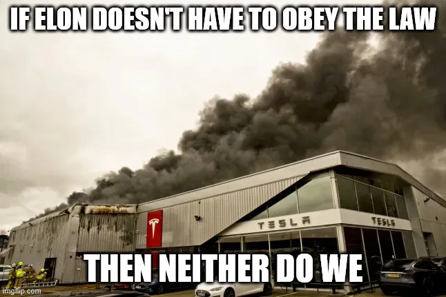 A sepia-toned photo shows a large industrial building with 'TESLA' signage and a red Tesla logo, engulfed in thick black…