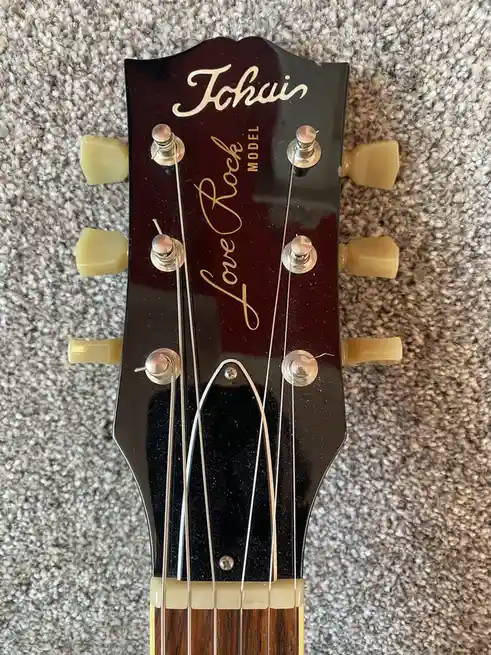 A close-up photo of a guitar headstock with the brand name 'Johari' and the model name 'Love Rock' written in gold cursive…