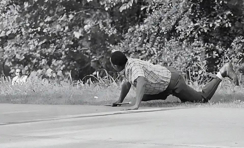 A black and white photo shows a man in a plaid shirt and trousers lying prone on a paved surface, propping himself up on his…