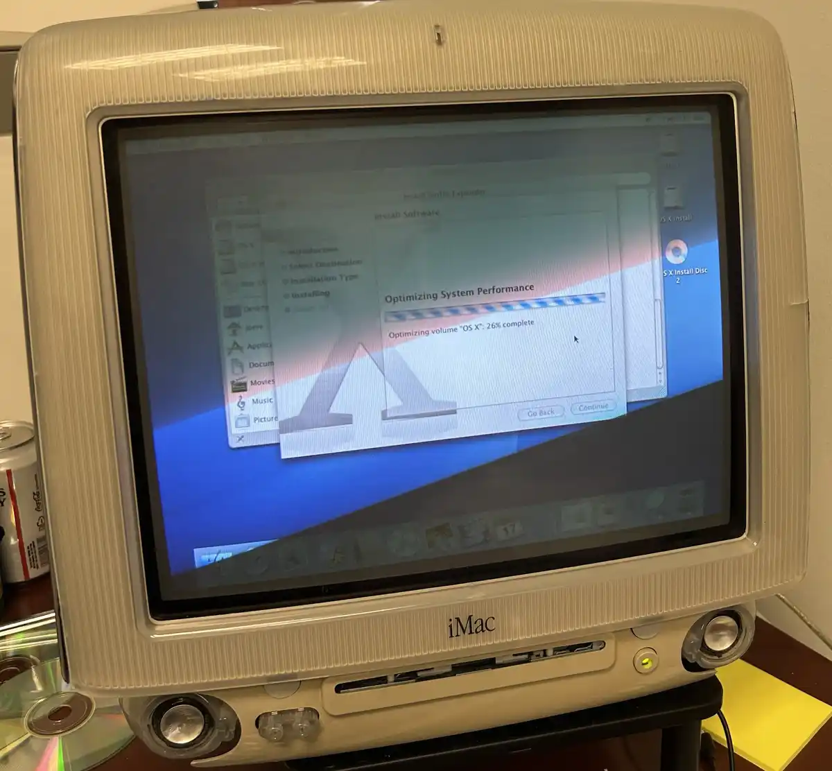 A vintage beige iMac G3 computer displays an OS X installation window showing 'Optimizing System Performance' at 26%…