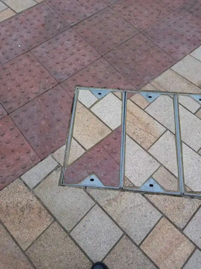 A close-up photo shows a metal manhole cover set into a tiled sidewalk, its surface patterned with beige and red tiles…