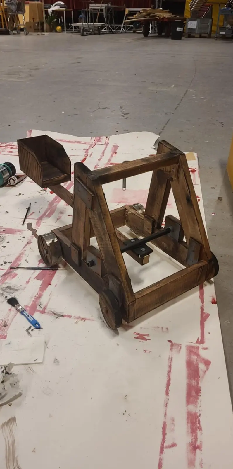 A small, wooden trebuchet sits on a white sheet with red paint splatters in a workshop. The machine has a bucket-like…