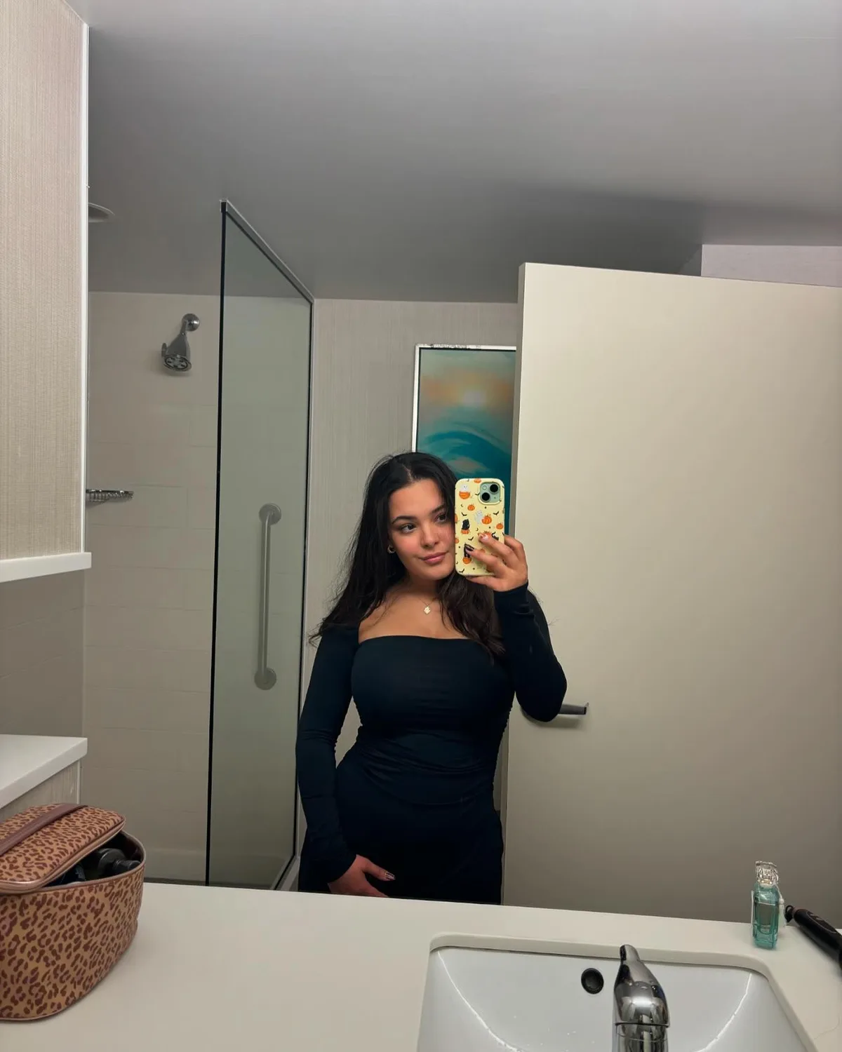 A young woman with long dark hair takes a mirror selfie in a modern bathroom, wearing a black off-the-shoulder dress and…