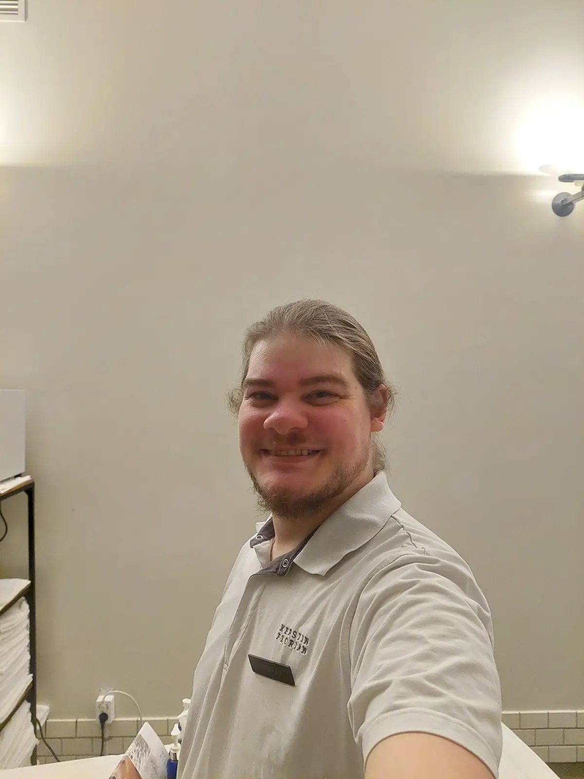 A man with long hair and a beard, wearing a light-colored polo shirt with a name tag reading 'KERSTIN FLORIAN', smiles at…