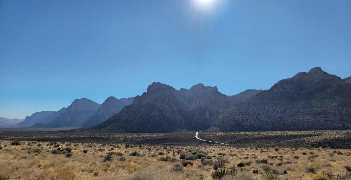 A wide desert landscape under a bright sun, featuring rugged mountains in the distance and a winding road cutting through…