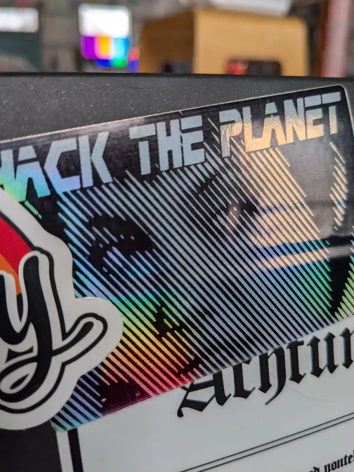A close-up of a holographic sticker on a black surface that reads 'HACK THE PLANET' in bold white letters, with diagonal…