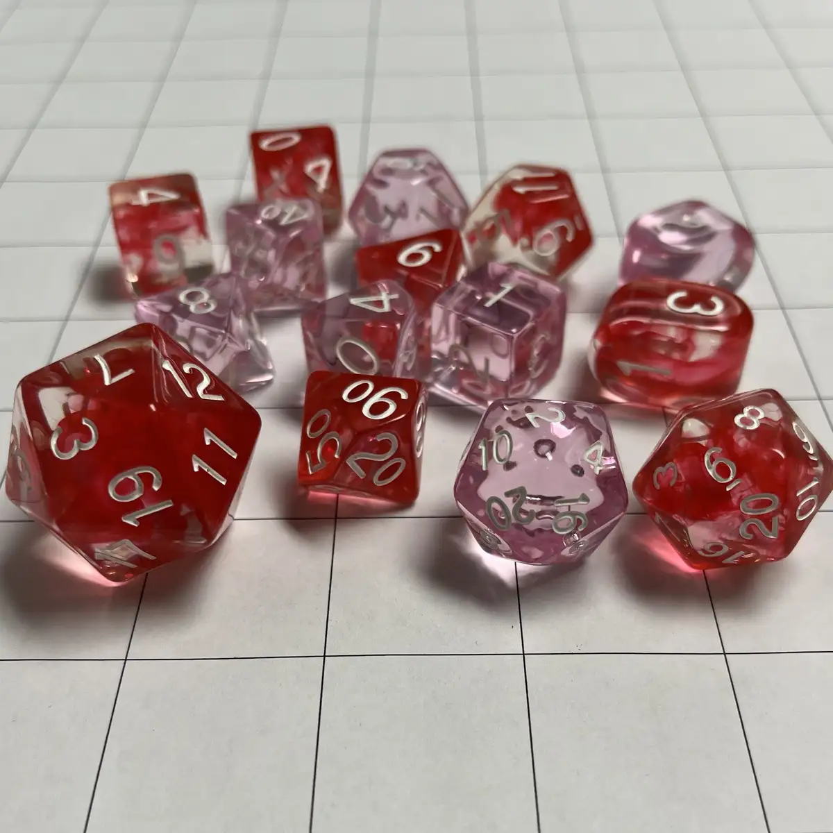 A collection of translucent red and pink polyhedral dice with silver numbers are scattered on a white gridded surface, all…