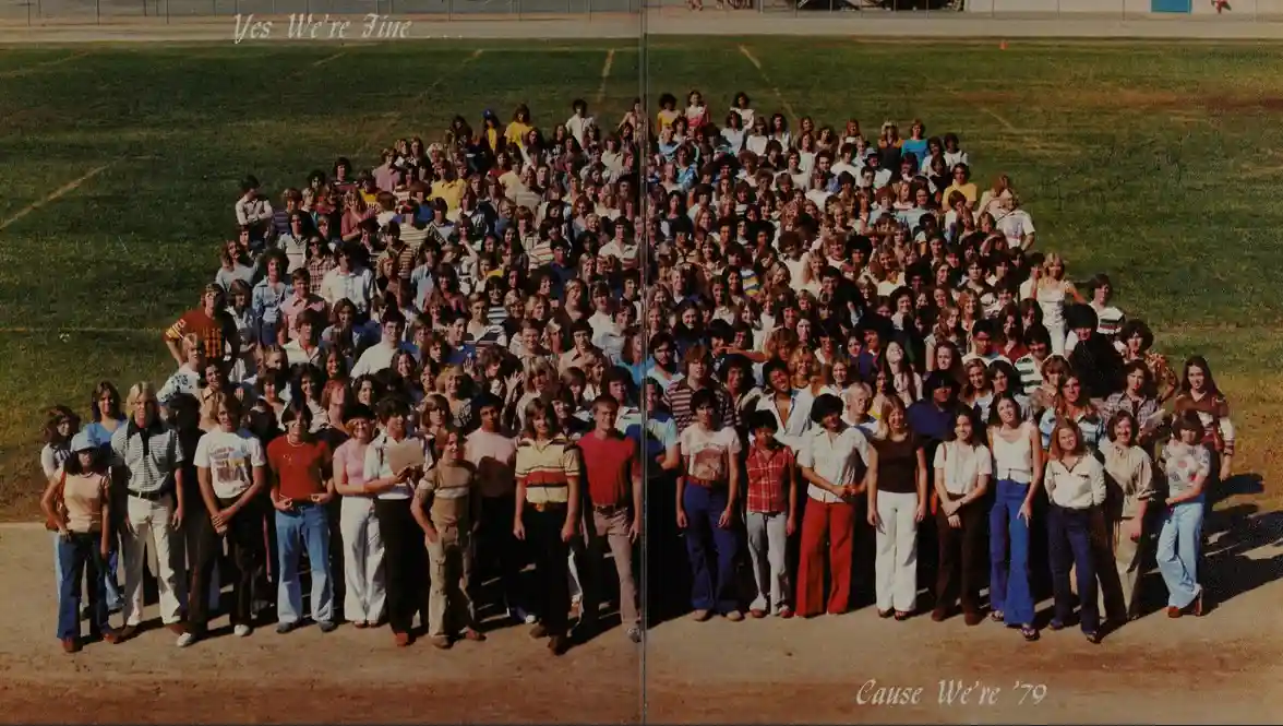 A vintage school photo of a large group of students and staff arranged in rows on a grassy field, with the text 'Yes We're…