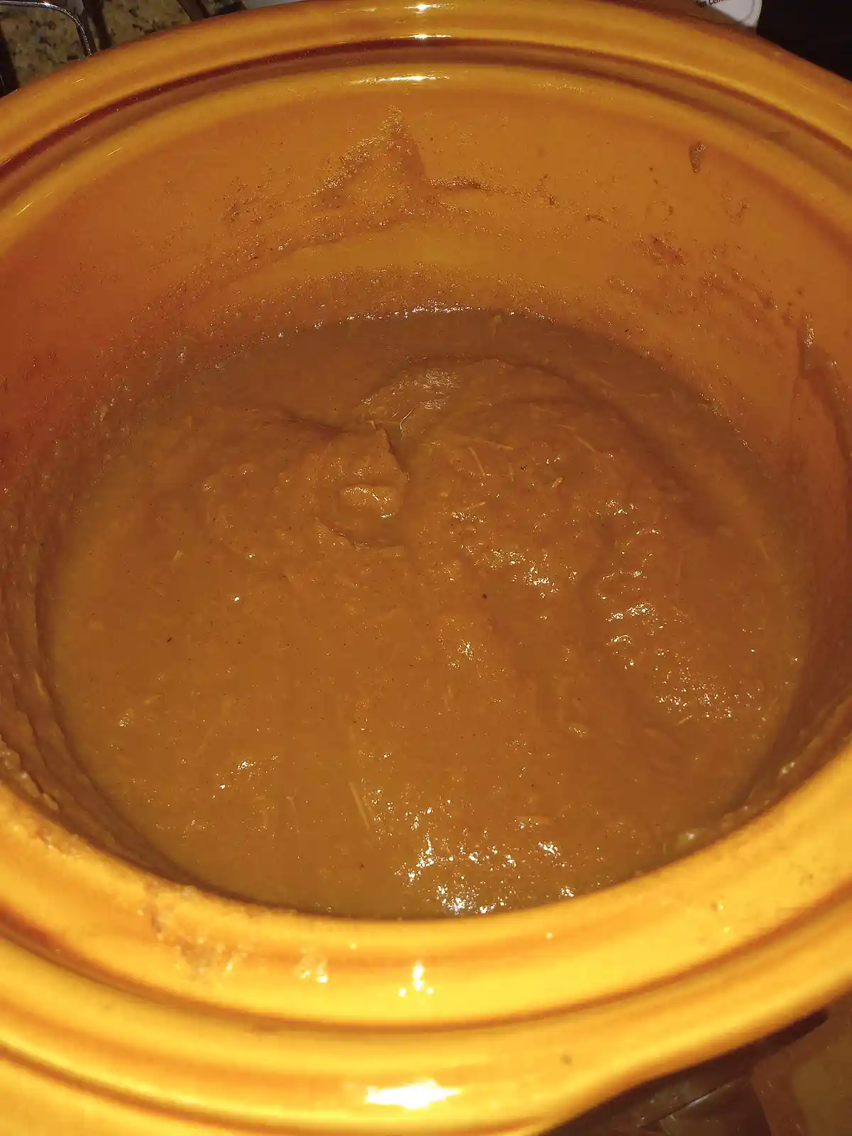 A close-up photo shows the inside of a yellow ceramic crockpot filled with a thick, brown, soupy liquid that appears mostly…