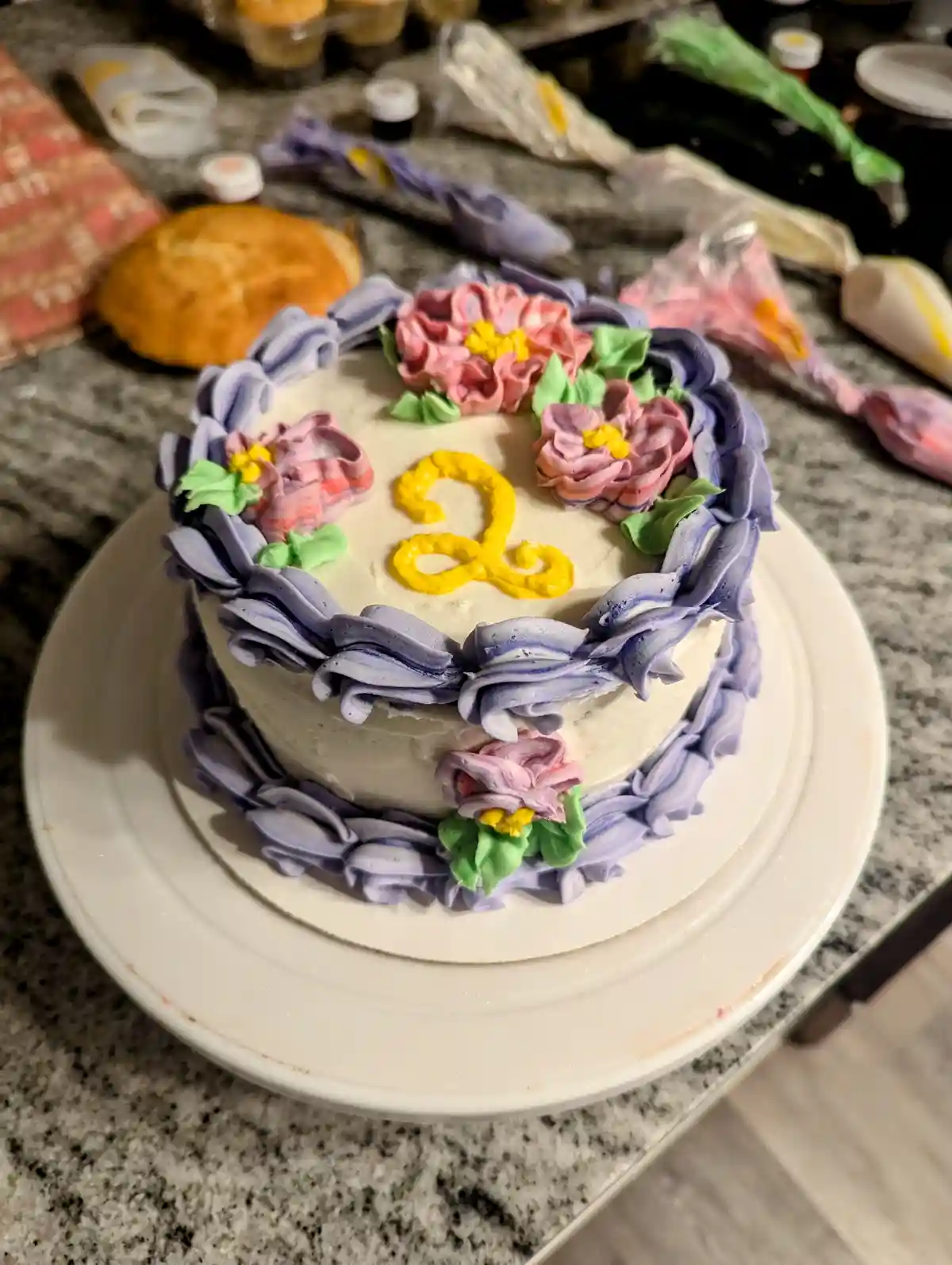 A round cake with white frosting sits on a white plate on a granite countertop, decorated with purple ruffled borders, pink…