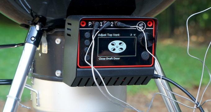 A close-up of a digital control panel attached to a grill, showing a screen with a film reel icon and the text 'Adjust Top…
