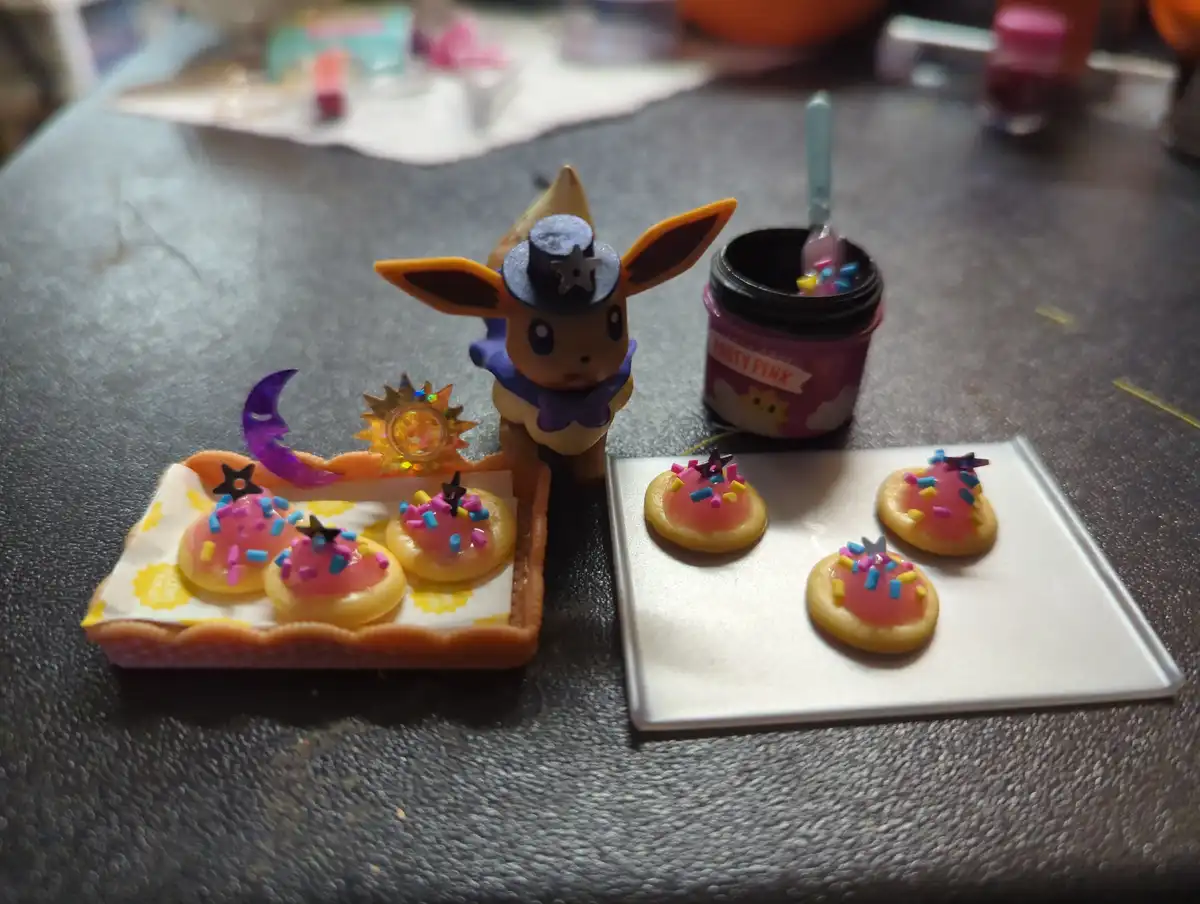 A small Eevee figurine wearing a police hat and purple bowtie stands beside two trays of miniature cookies decorated with…