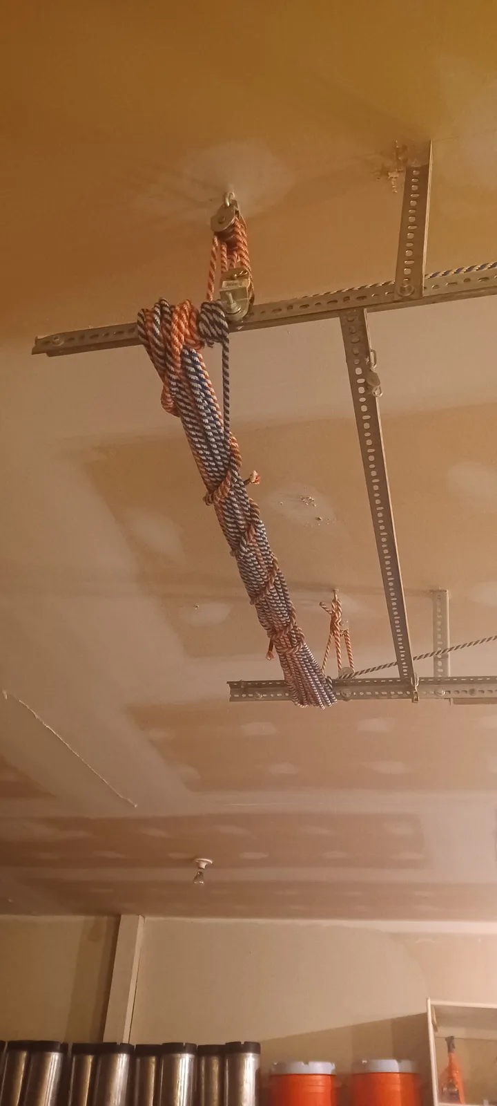 A photo shows a complex rope and pulley system rigged between metal beams attached to a ceiling, with thick ropes hanging…